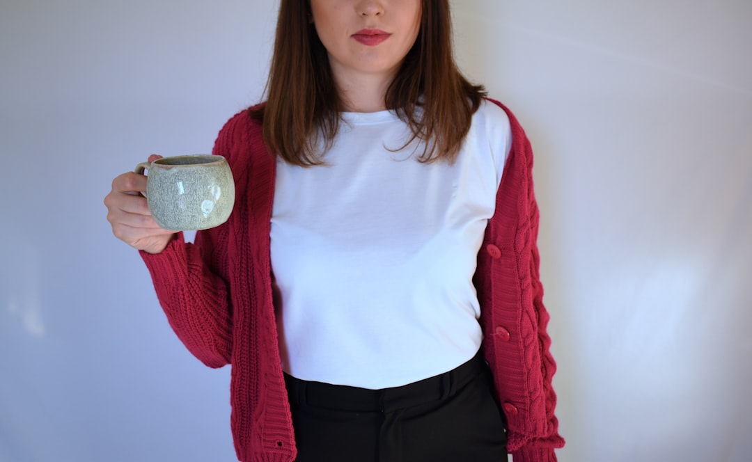 woman holding a cup of coffee at right hand and reading book on her lap while holding it open with her left hand in a well-lit room